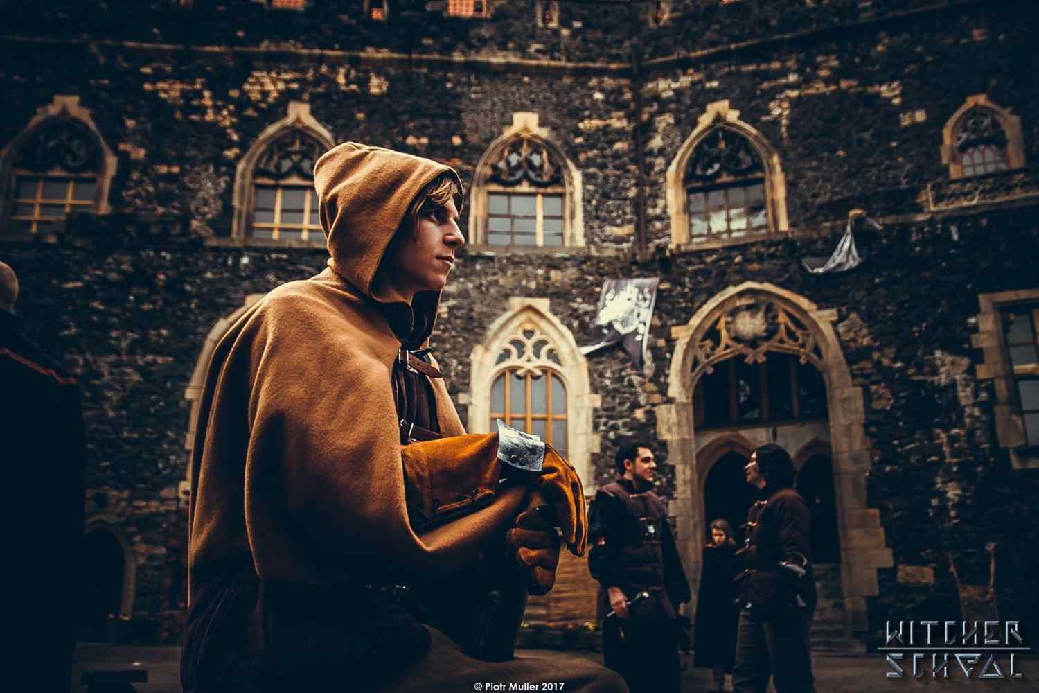 A cosplayer dressed in medieval-style robes at the Witcher School event in Poland, standing in front of a gothic-style stone castle, surrounded by other participants in costume.