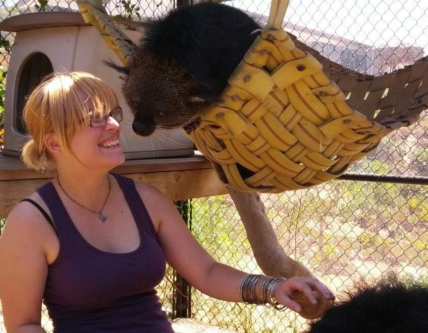 Timbre Webb smiling and interacting with a binturong lounging in a woven hammock at an animal sanctuary, enjoying a moment of connection.